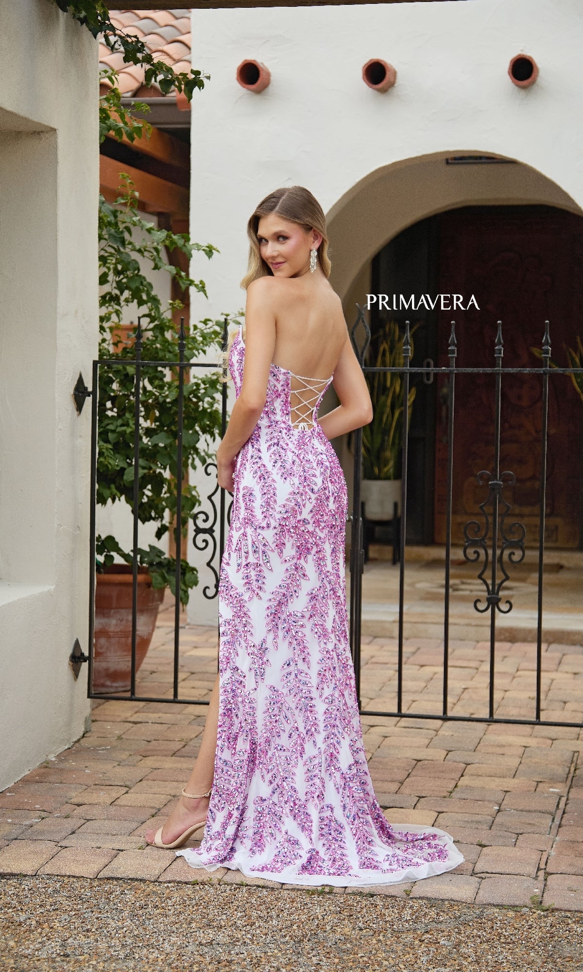 A woman stands outdoors in front of a wrought iron gate and Mediterranean-style building wearing the strapless Primavera 4314 long sequin prom dress in white and pink, featuring an open lace-up back.
